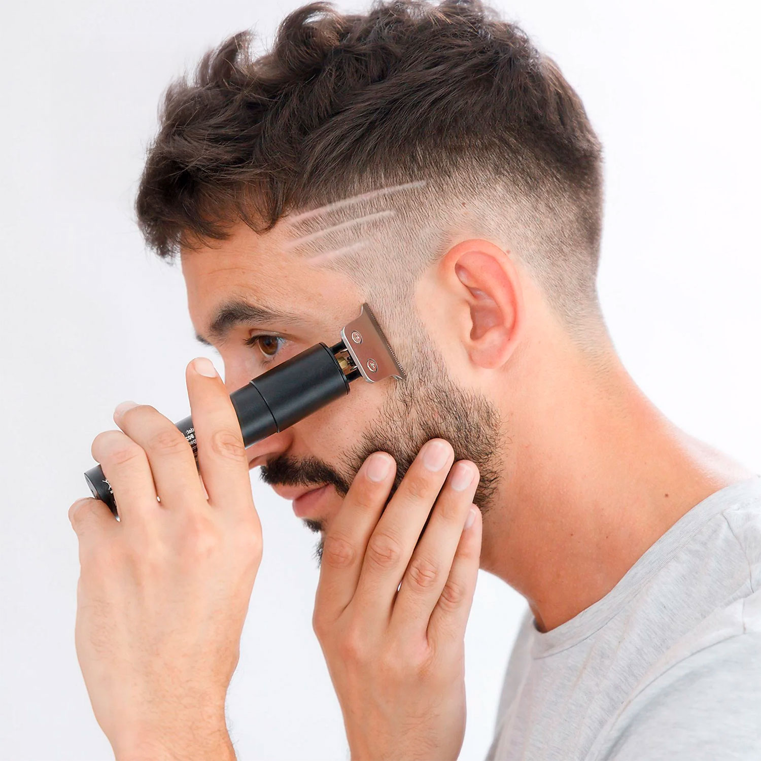 Un hombre se afeita el pelo con una recortadora eléctrica. Tiene barba y el fondo es blanco.