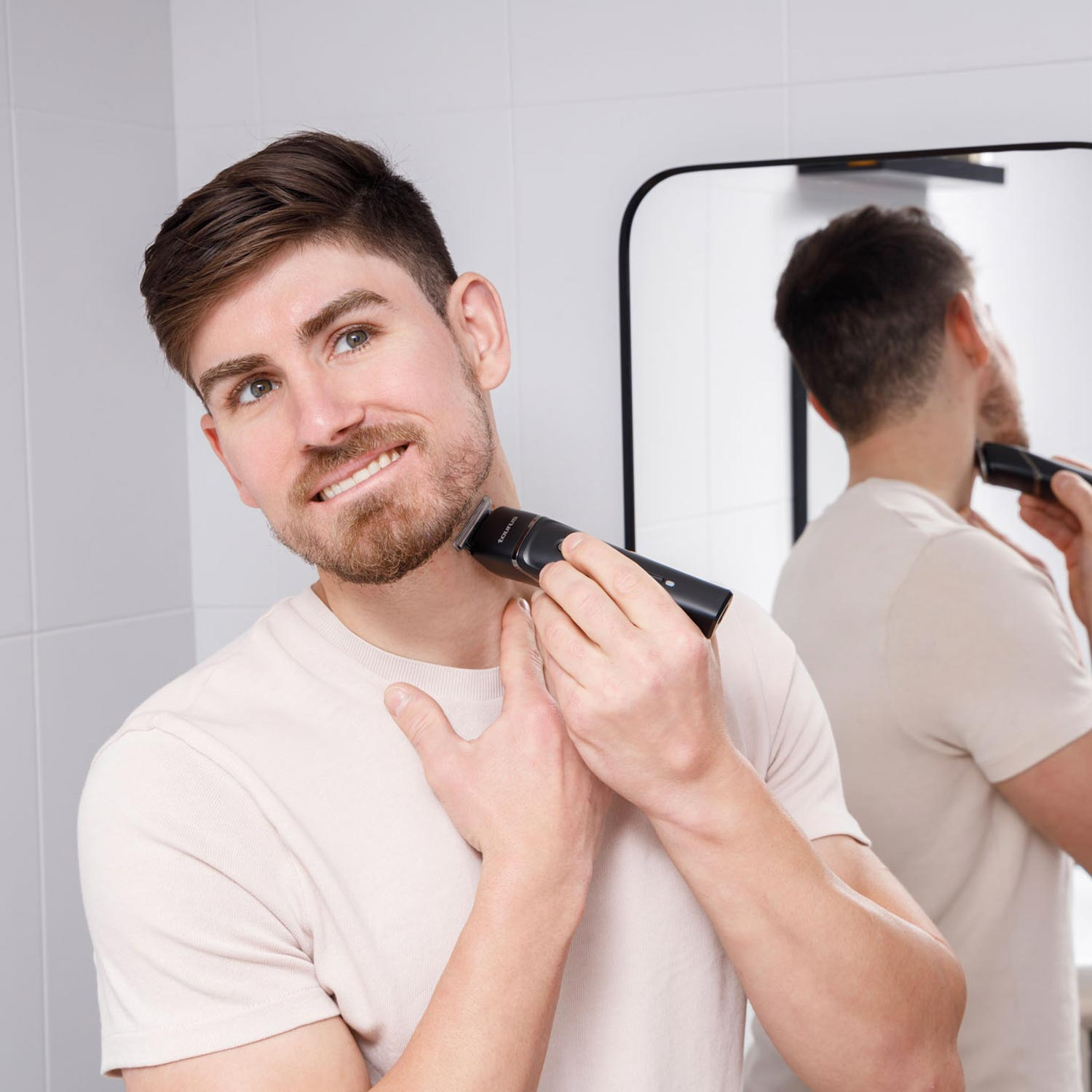 Un hombre se afeita el cuello en un baño. Sostiene una recortadora negra, mirando al espejo. También es visible su reflejo.