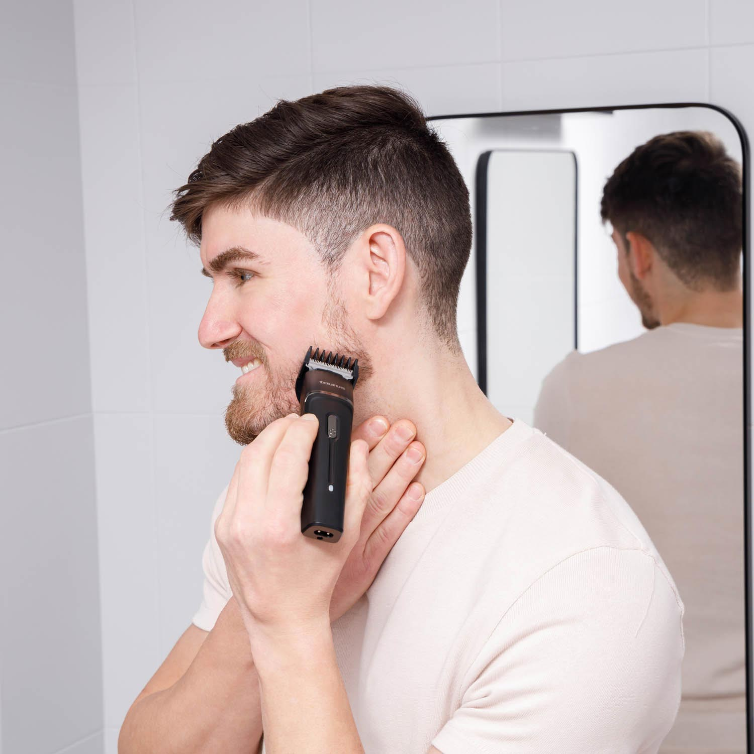 Un hombre se afeita la barba con una recortadora eléctrica frente a un espejo, vistiendo una camisa de color claro en un baño.