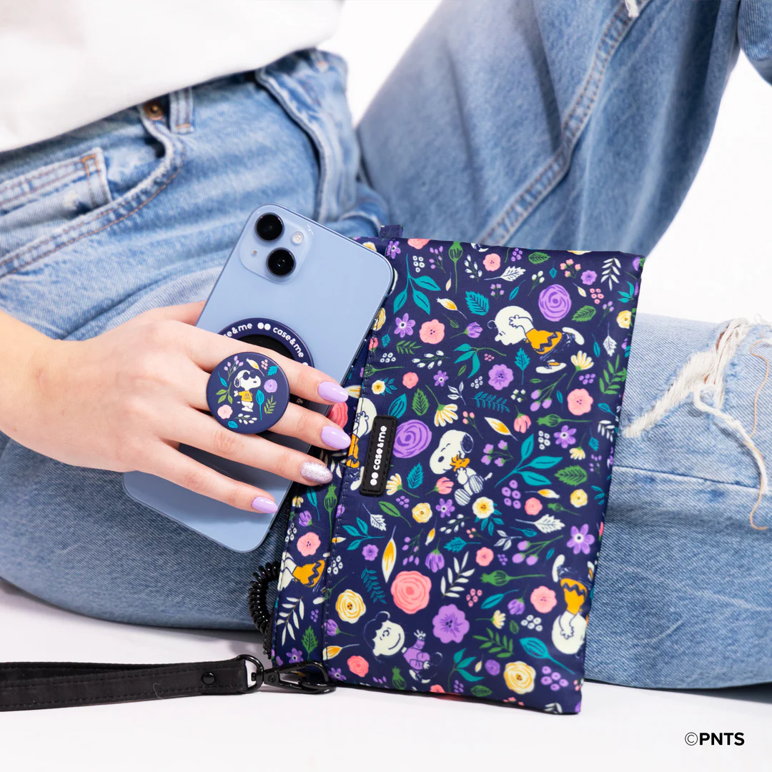 Osoba trzyma niebieski telefon z popsocketem Snoopy obok kwiatowego portfela. Siedzi w dżinsach na białym tle.