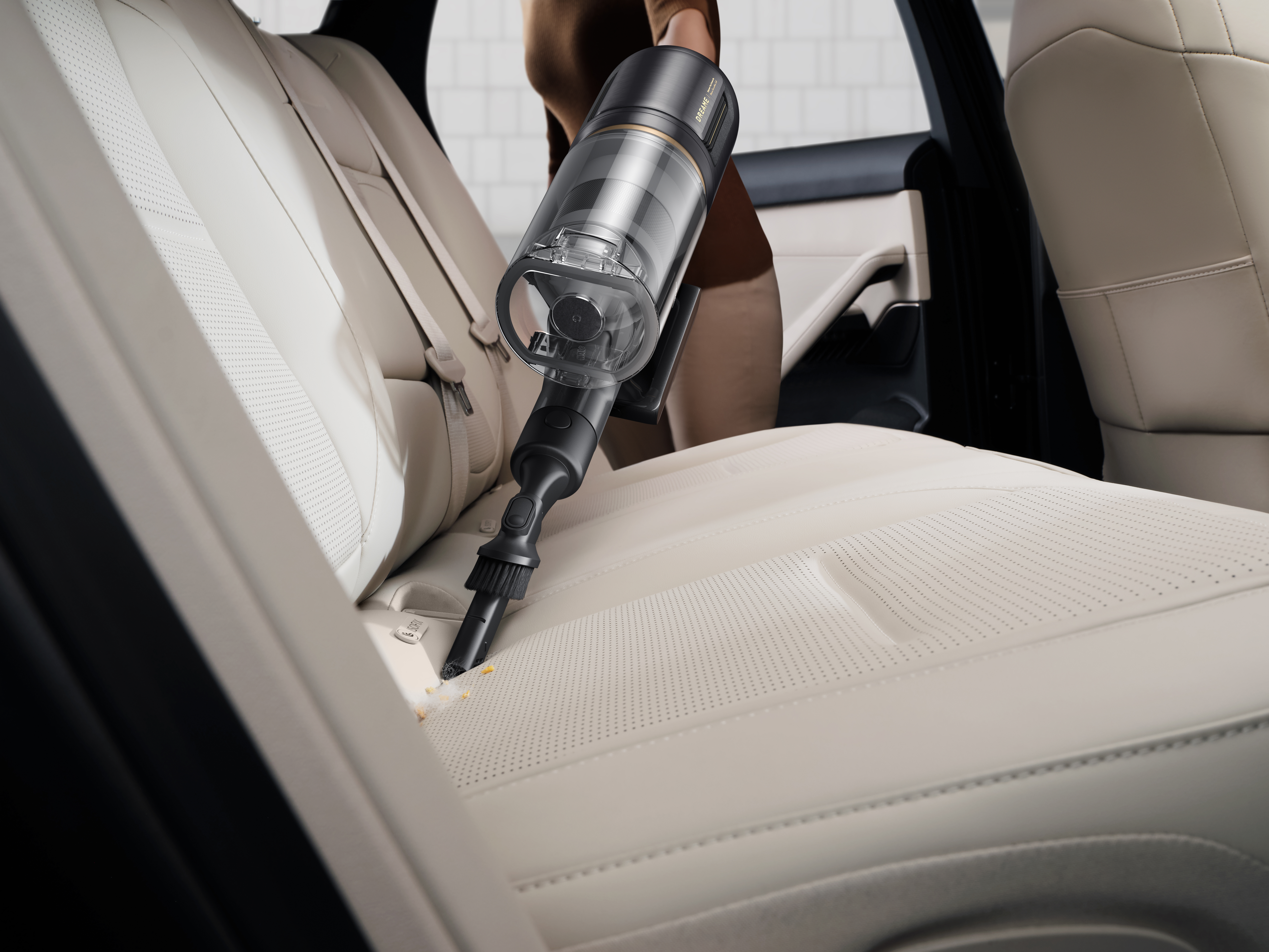 Osoba odkurza siedzenie w samochodzie odkurzaczem ręcznym. Siedzenie jest kremowe z perforacjami.