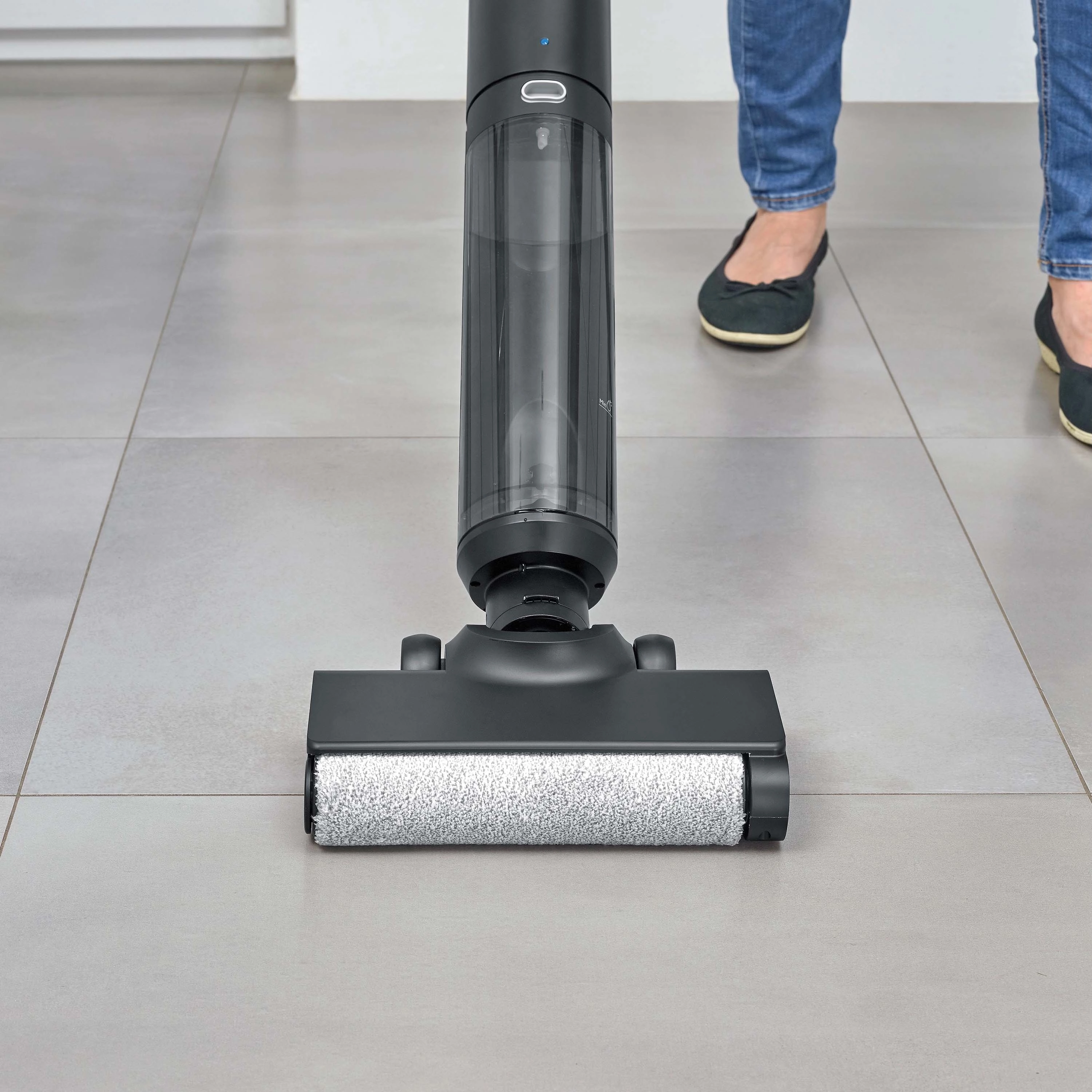 Osoba używa czarnego środka do czyszczenia podłóg na szarej, kafelkowej podłodze. Środek czyszczący ma zbiornik na wodę. Osoba nosi dżinsy i czarne baleriny.