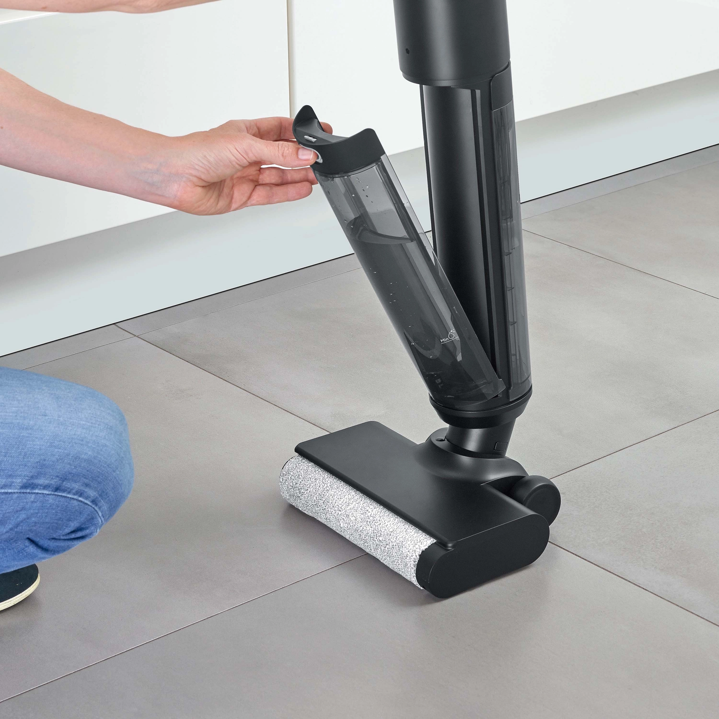 Osoba trzyma zbiornik na wodę czarnej myjki do podłóg. Widoczna jest część nóg osoby.