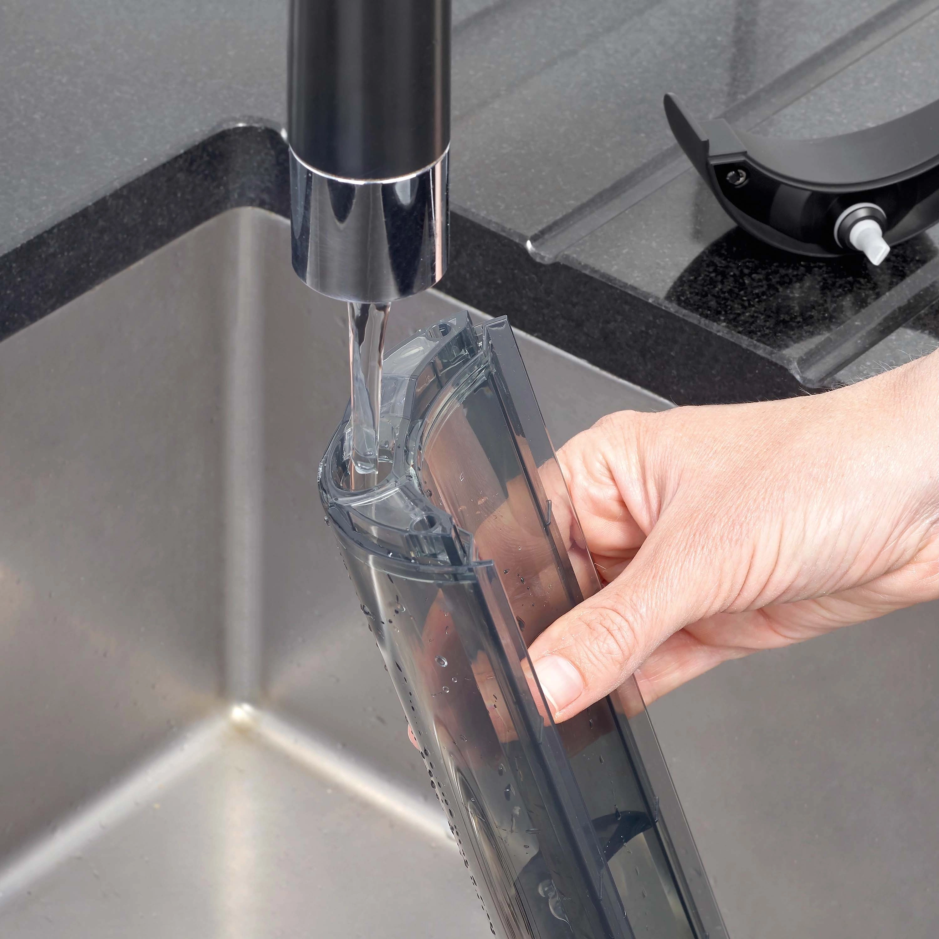 Ręka trzyma zbiornik na wodę pod kranem, który napełnia się wodą. Zlewozmywak i blat są ciemnoszare.