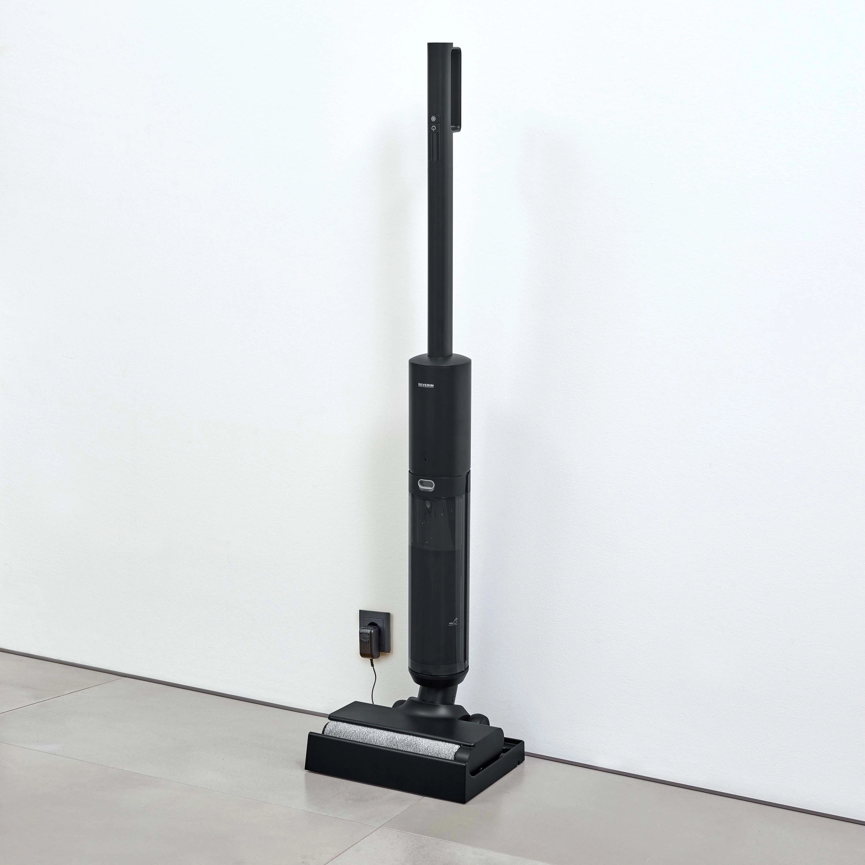 Czarny odkurzacz pionowy stoi przy białej ścianie. Stacja ładująca jest podłączona do gniazdka w pobliżu.