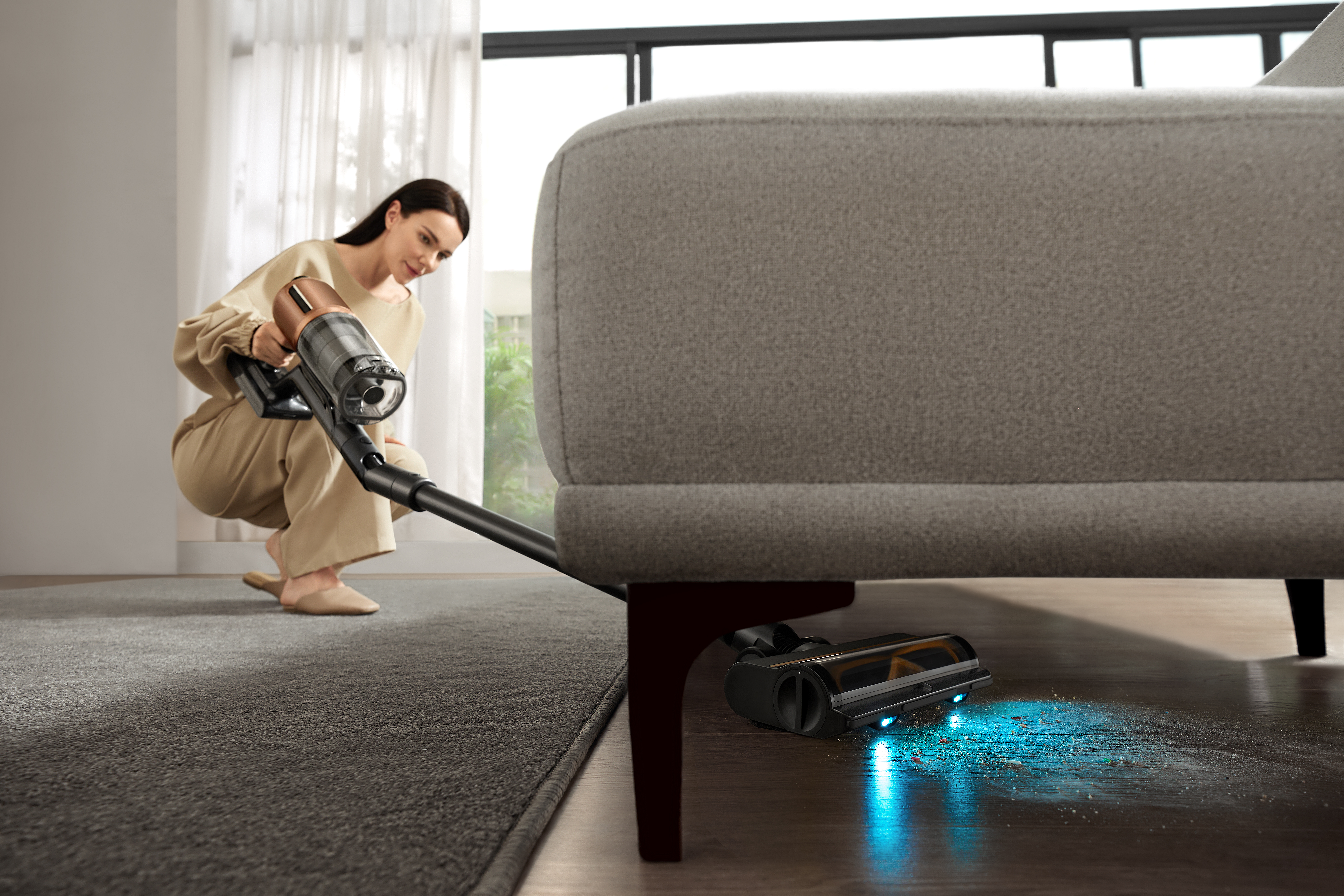 Kobieta odkurza pod kanapą. Używany jest czarny i brązowy odkurzacz. Podłoga jest drewniana.
