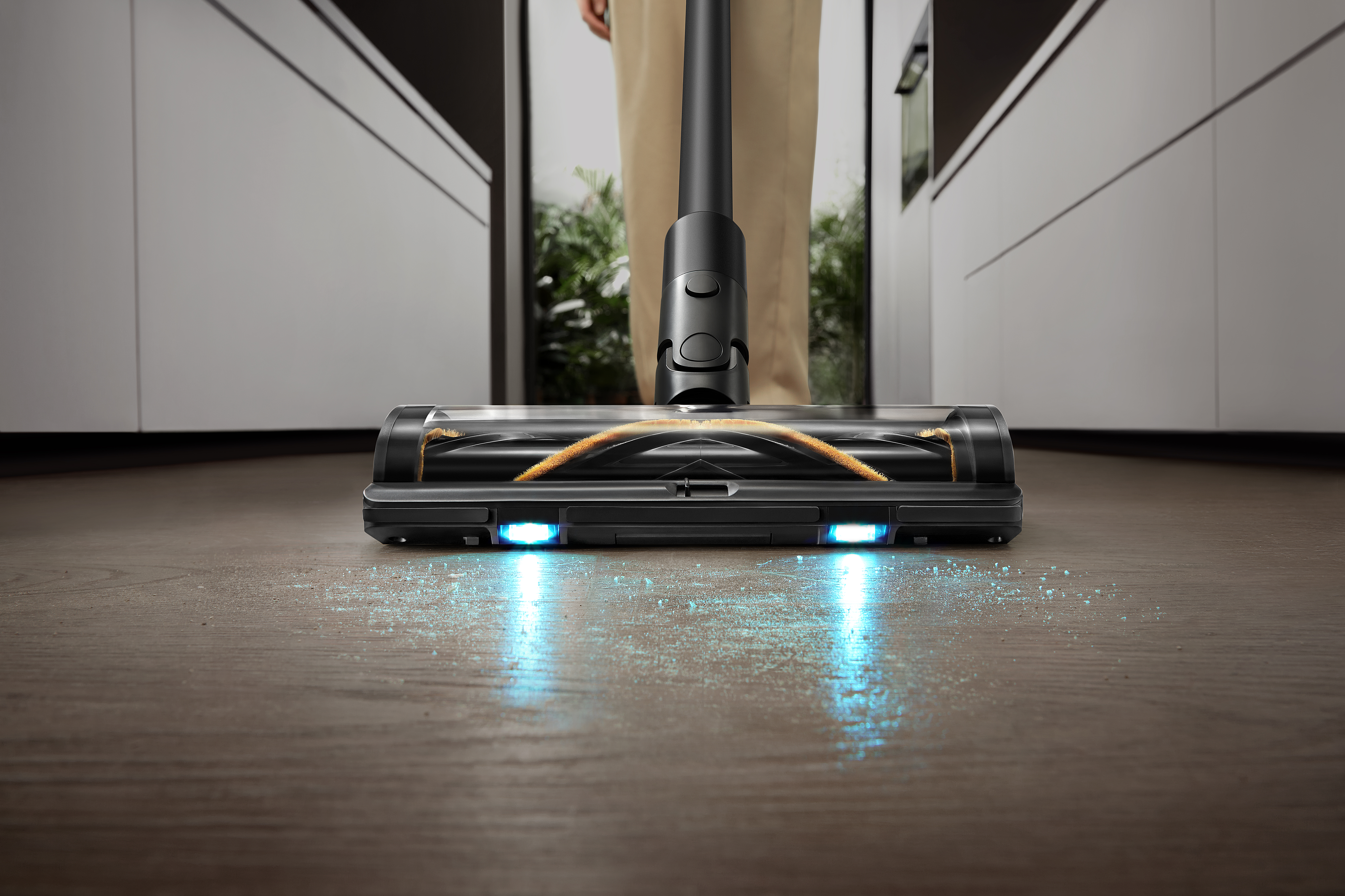 Czarna głowica odkurzacza na drewnianej podłodze. Dwa niebieskie światła są włączone. Osoba stoi za odkurzaczem.