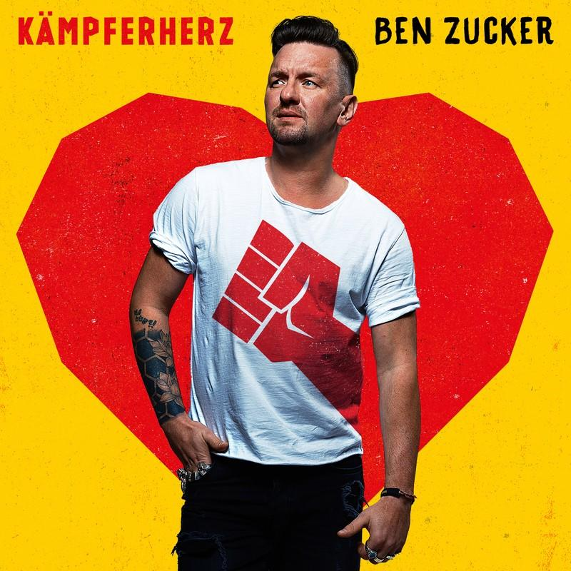 Mann in weißem T-Shirt mit rotem Faust-Design steht vor rotem Herz auf gelbem Hintergrund. Text: KÄMPFERHERZ und BEN ZUCKER.