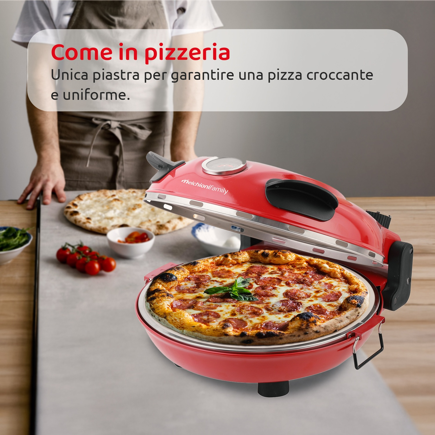 Un forno per pizza rosso con una pizza cotta all'interno. Una persona con un grembiule dietro un tavolo con ingredienti.