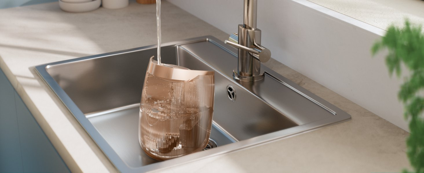 El agua se vierte en un filtro de agua de vidrio en un fregadero de acero inoxidable. Hay un grifo a la derecha y plantas verdes a la derecha.