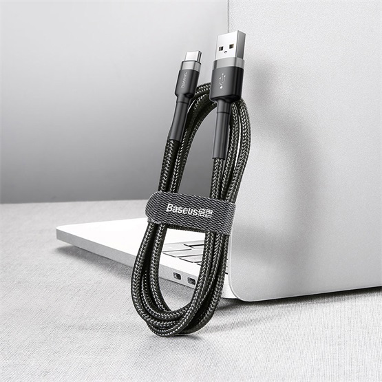 Fekete és szürke USB-C kábel USB porttal. A kábelt egy tépőzáras pánt tartja. A háttérben egy laptop látható.