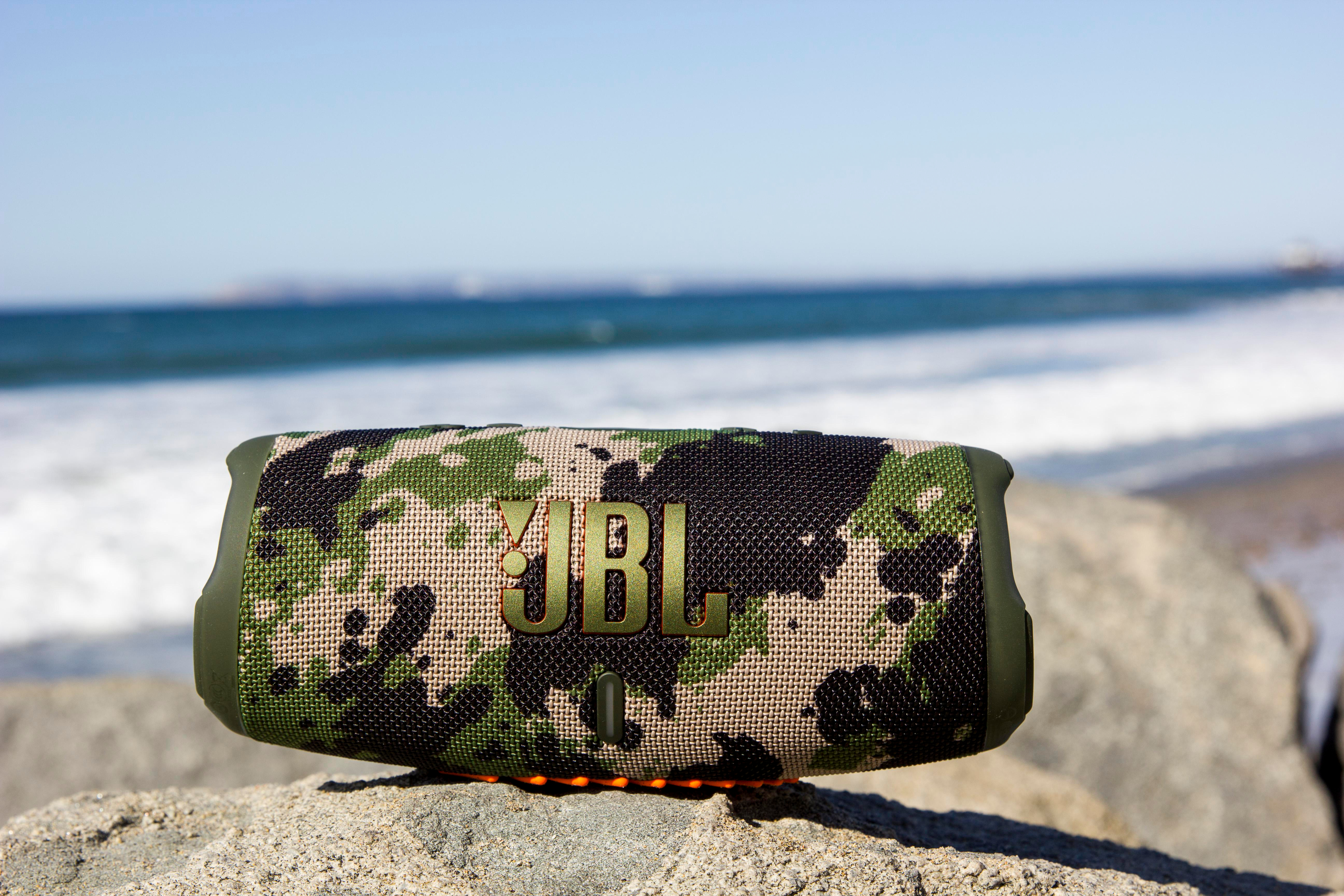 Ein tarnfarbener JBL-Lautsprecher liegt auf einem Felsen, mit Meer und Himmel im Hintergrund.