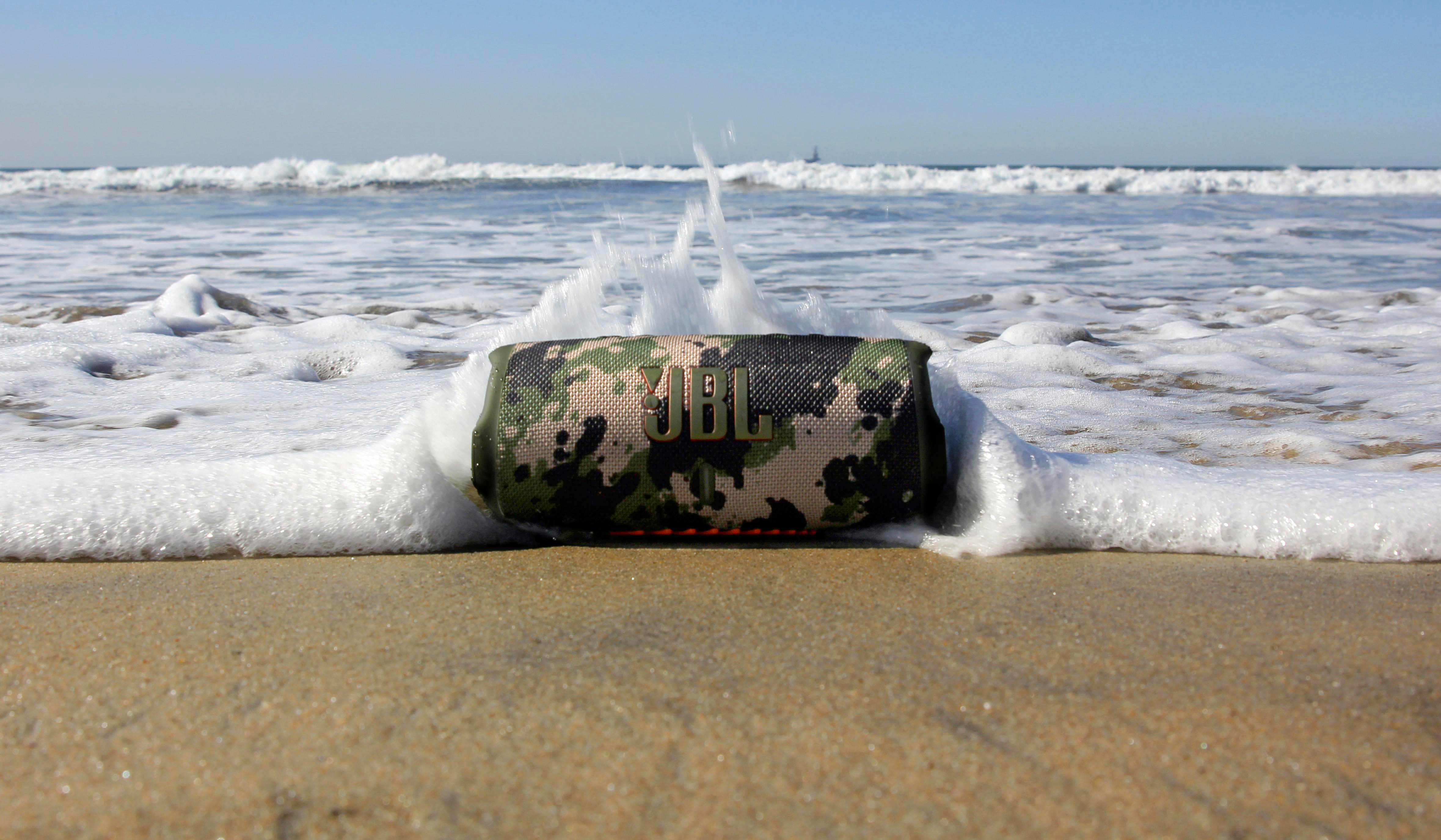Ein Camouflage-JBL-Lautsprecher an einem Sandstrand, der von einer Welle getroffen wird. Der Ozean ist im Hintergrund unter blauem Himmel.