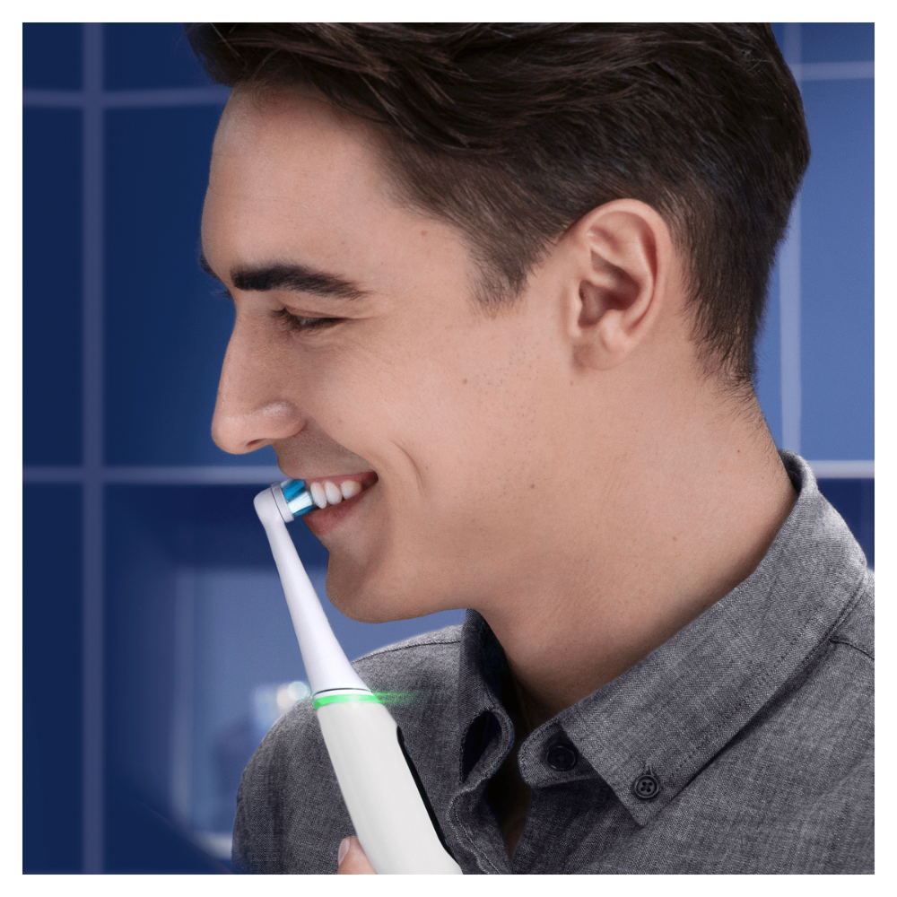 Un hombre sonríe mientras se cepilla los dientes con un cepillo eléctrico. Viste una camisa gris. El fondo tiene azulejos azules.