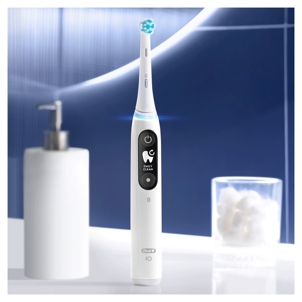 Cepillo de dientes eléctrico Oral-B iO blanco, con cabezal azul y blanco, junto a un dispensador de jabón y un vaso con bolas de algodón.