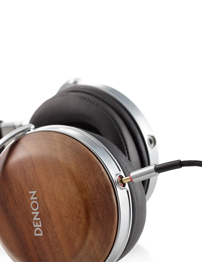 Zbliżenie słuchawek z drewnianymi nausznikami i srebrnymi wykończeniami. Wpięty jest kabel. Widoczna jest nazwa marki DENON.