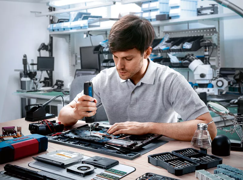 Egy férfi laptopot javít egy eszközzel. Különféle elektronikai alkatrészek és szerszámok vannak az asztalon.