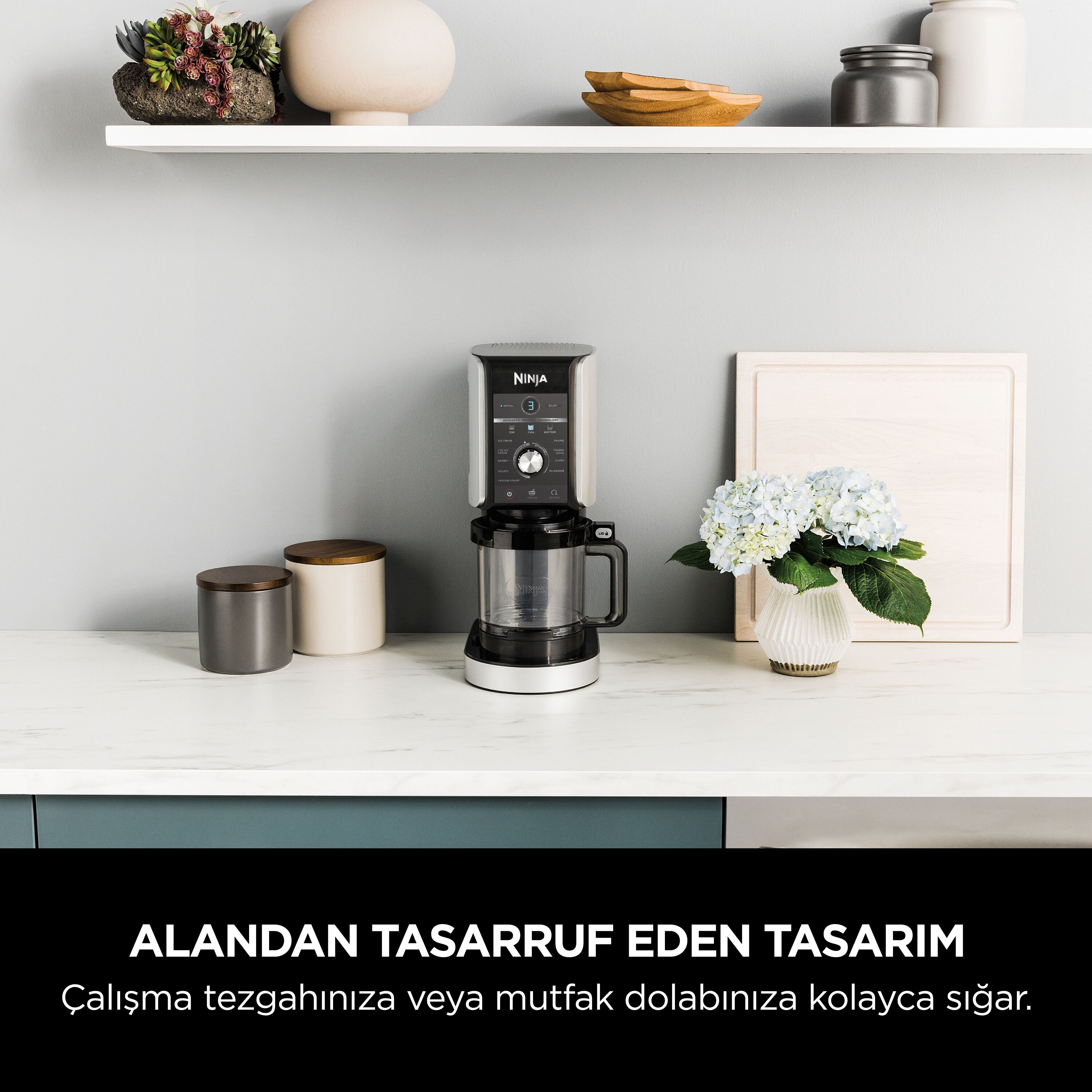 Bir Ninja mutfak aleti tezgahta. Bir vazoda çiçekler ve kaplar tezgahta. Üstte bir raf var.