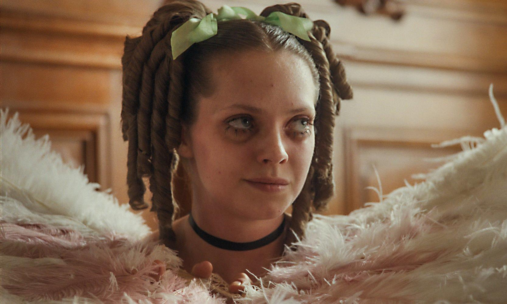 Eine junge Frau mit Locken und grünen Schleifen blickt zur Seite. Sie trägt einen Choker und ein Federkleid.