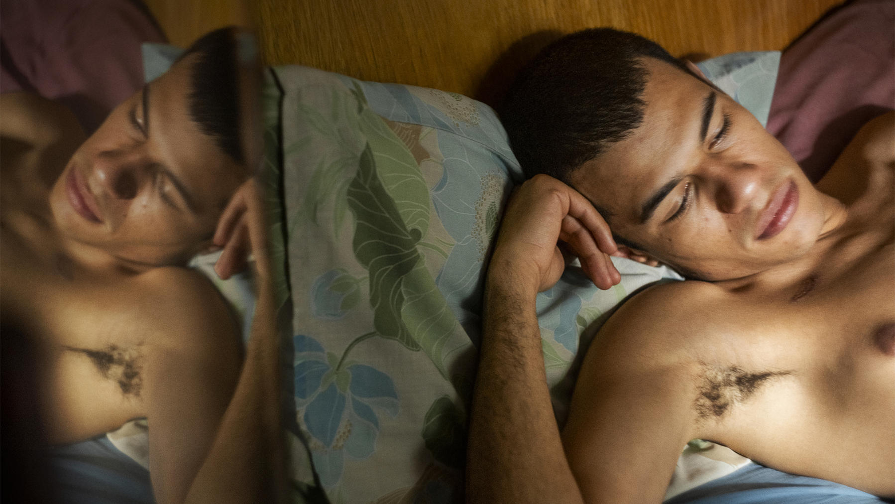 Ein Mann liegt mit geschlossenen Augen auf dem Bett, ohne Hemd. Sein Spiegelbild ist sichtbar. Er ruht auf einem Kissen mit Blumenmustern.