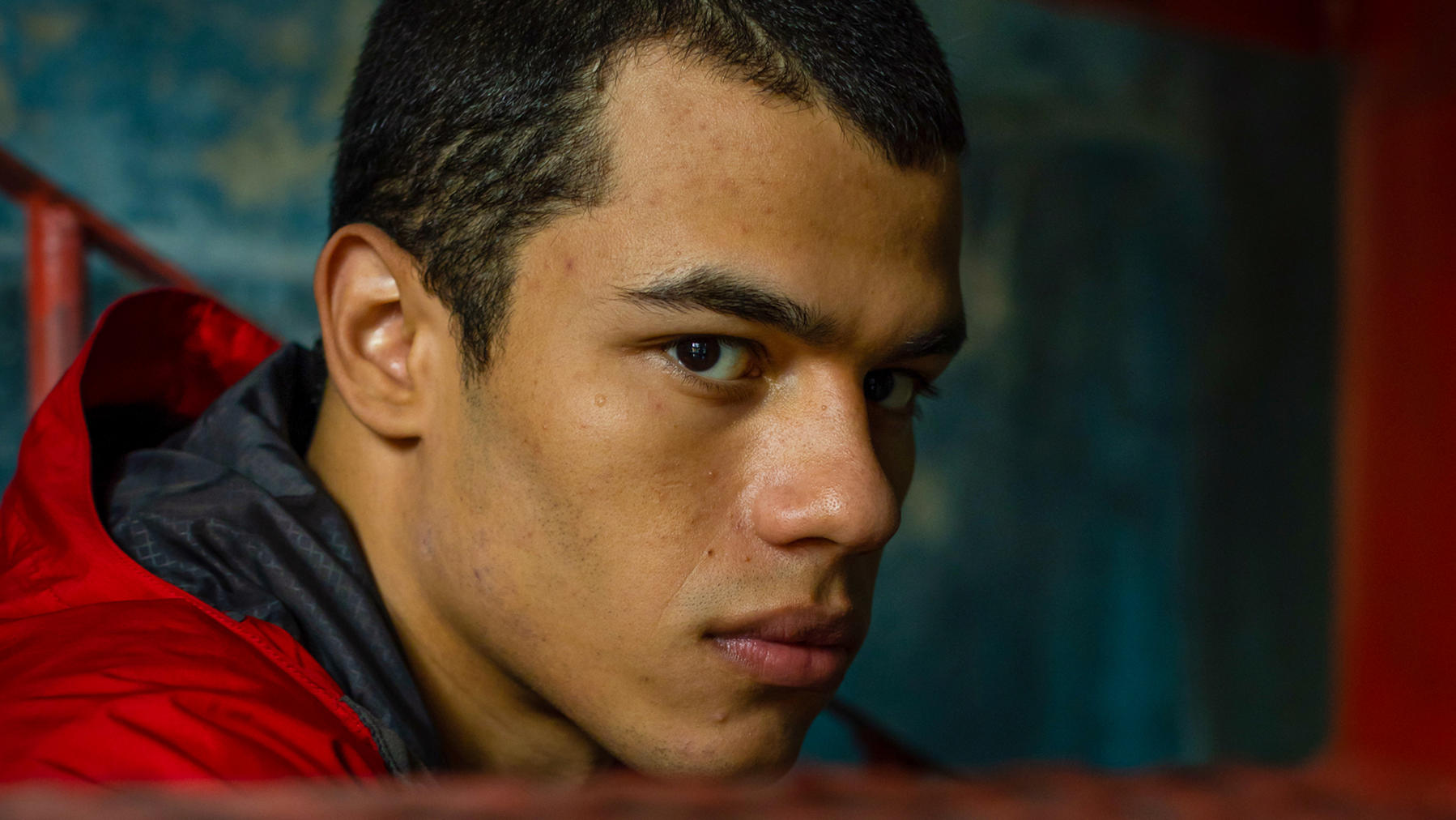 Nahaufnahme des Gesichts eines jungen Mannes. Er trägt eine rote Jacke und blickt in die Kamera. Hintergrund ist blau und rot.