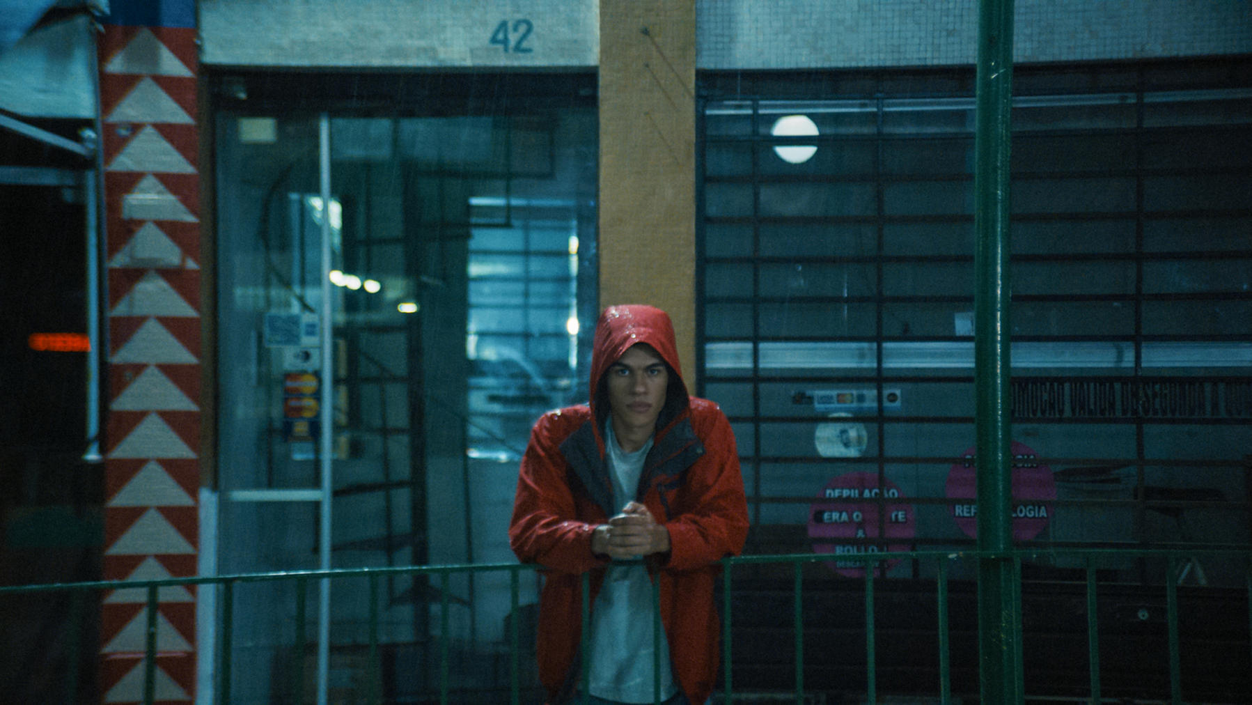 Ein junger Mann in einer roten Kapuzenjacke steht im Regen. Er steht vor einem Gebäude mit einem grünen Zaun.