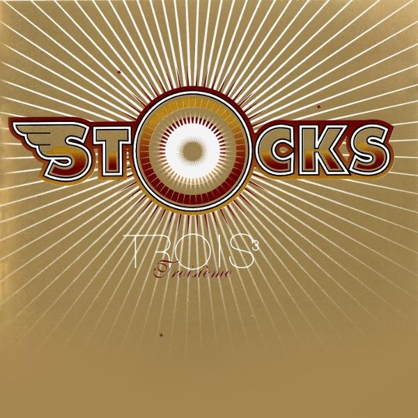 Goldener Hintergrund mit Strahlenlinien. Das Wort "STOCKS" über "TROIS" in einer stilisierten Schrift. In der Mitte ein Kreisdesign.