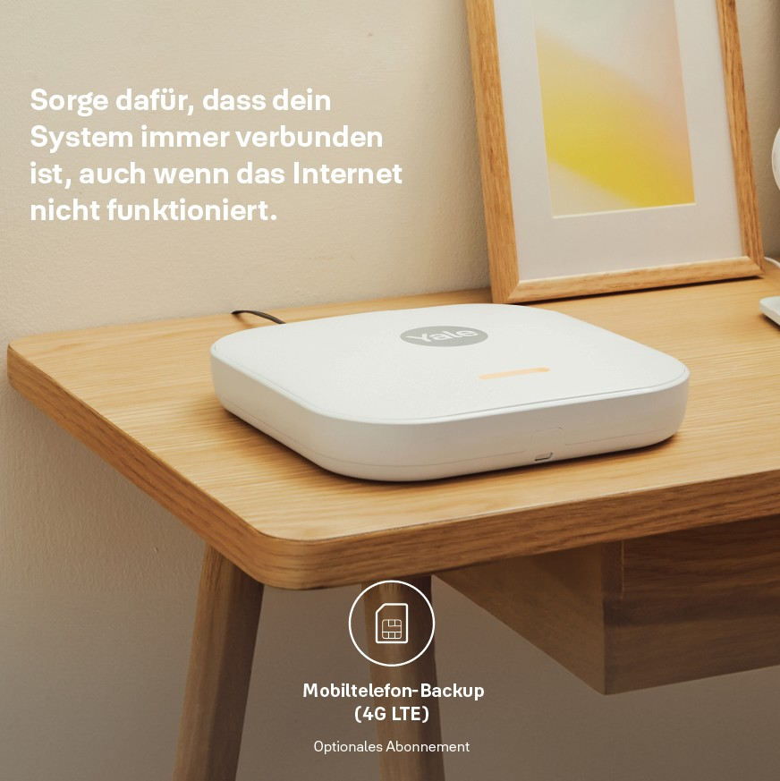Ein weißes Yale-Gerät auf einem Holztisch. Text-Overlay: Mobiltelefon-Backup (4G LTE). Ein gerahmtes Bild im Hintergrund.