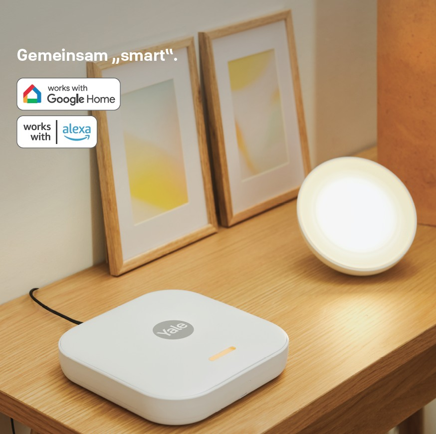 Ein weißes Yale-Gerät steht auf einem Holztisch. Eine leuchtende Lampe und zwei gerahmte Bilder stehen auf dem Tisch. Google Home und Alexa Icons sichtbar.