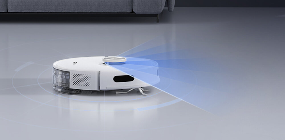 Biały robot odkurzający z niebieskim światłem, na szarej podłodze. Szara sofa w tle.