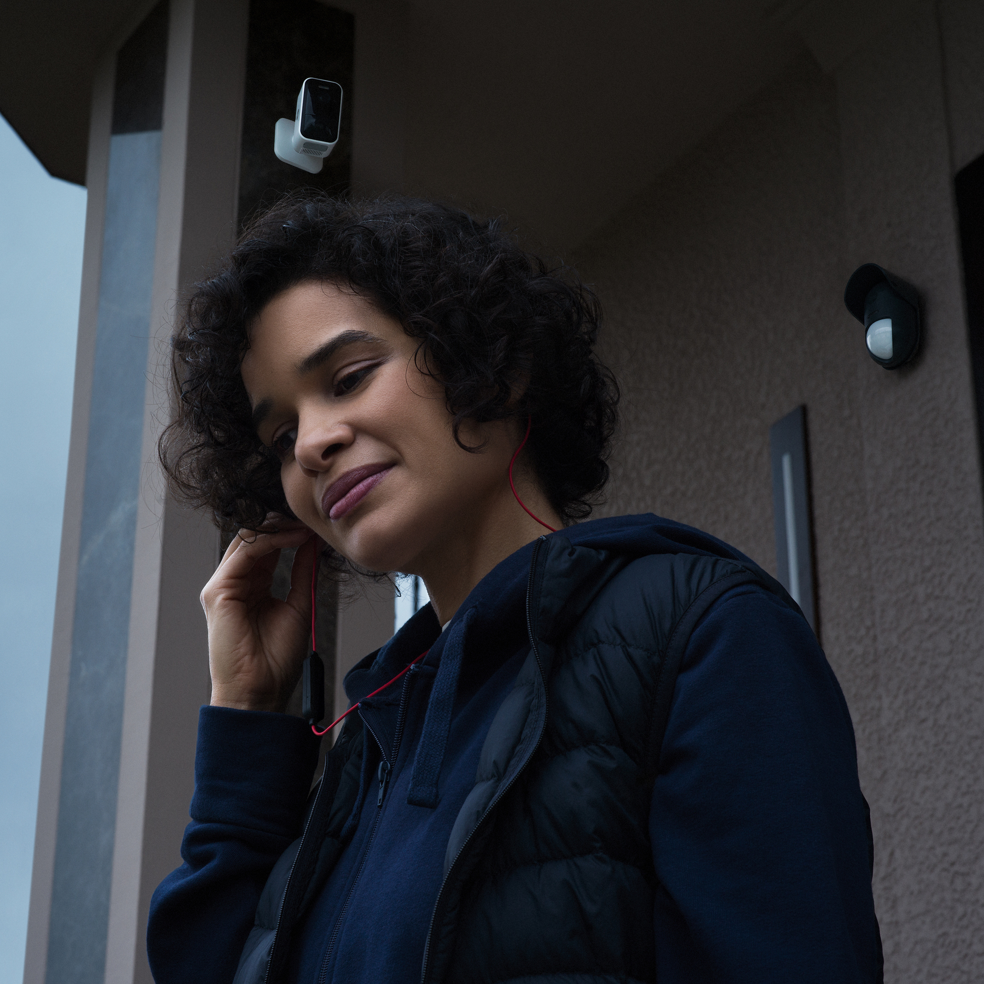 Frau, die rote Ohrhörer einstellt, trägt eine dunkle Jacke. Sicherheitskameras sind auf dem Gebäude hinter ihr zu sehen. Wolkiger Himmel.