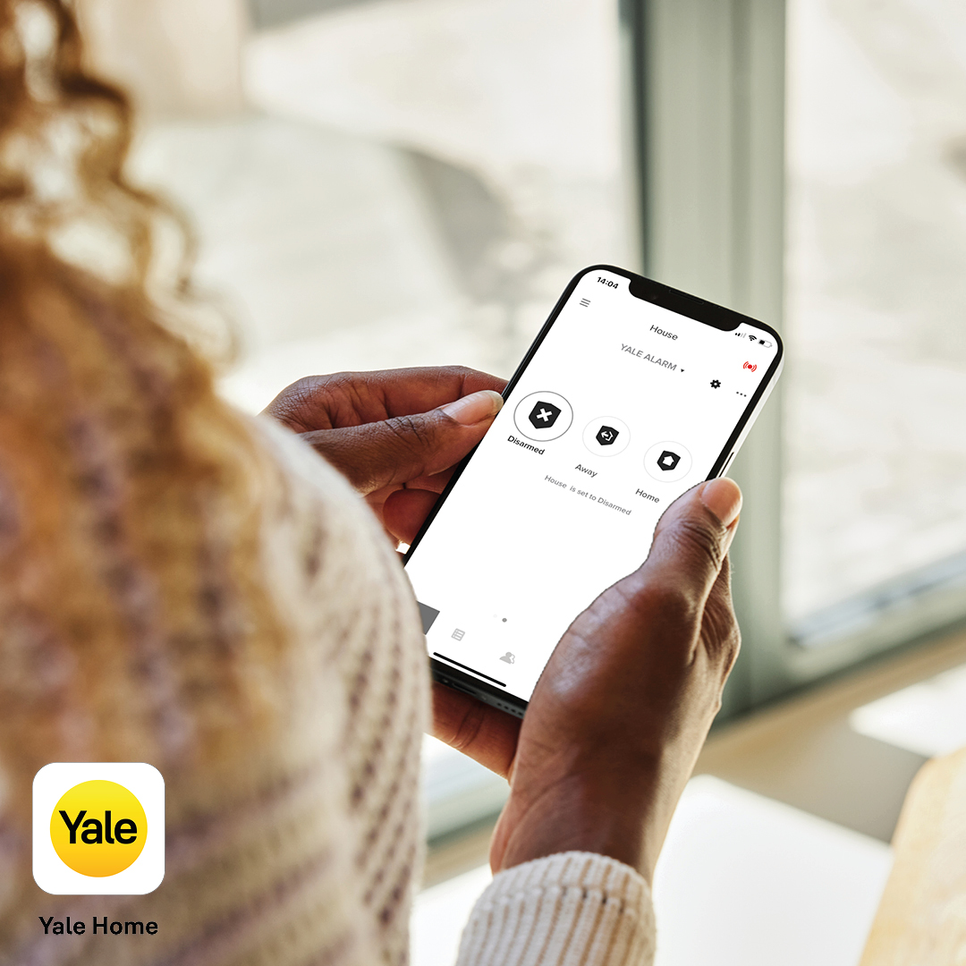 Person hält ein Telefon, das die Yale-Alarm-App zeigt. Der Bildschirm zeigt Optionen zum Deaktivieren, Scharfstellen und Aktivieren.