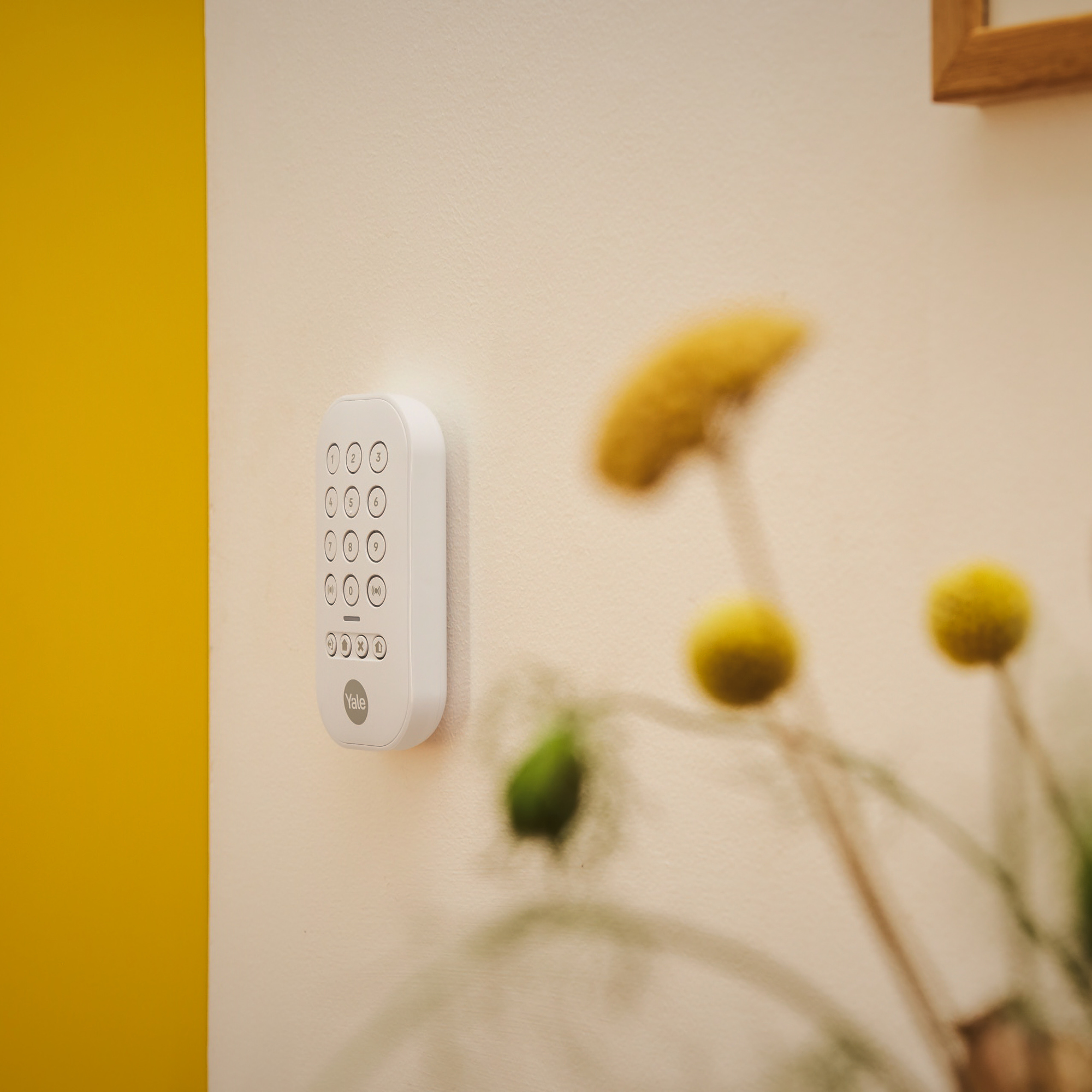 Ein weißes Yale-Tastenfeld an einer cremefarbenen Wand mit gelben Blumen im Hintergrund verschwommen. Gelber Wandabschnitt links.