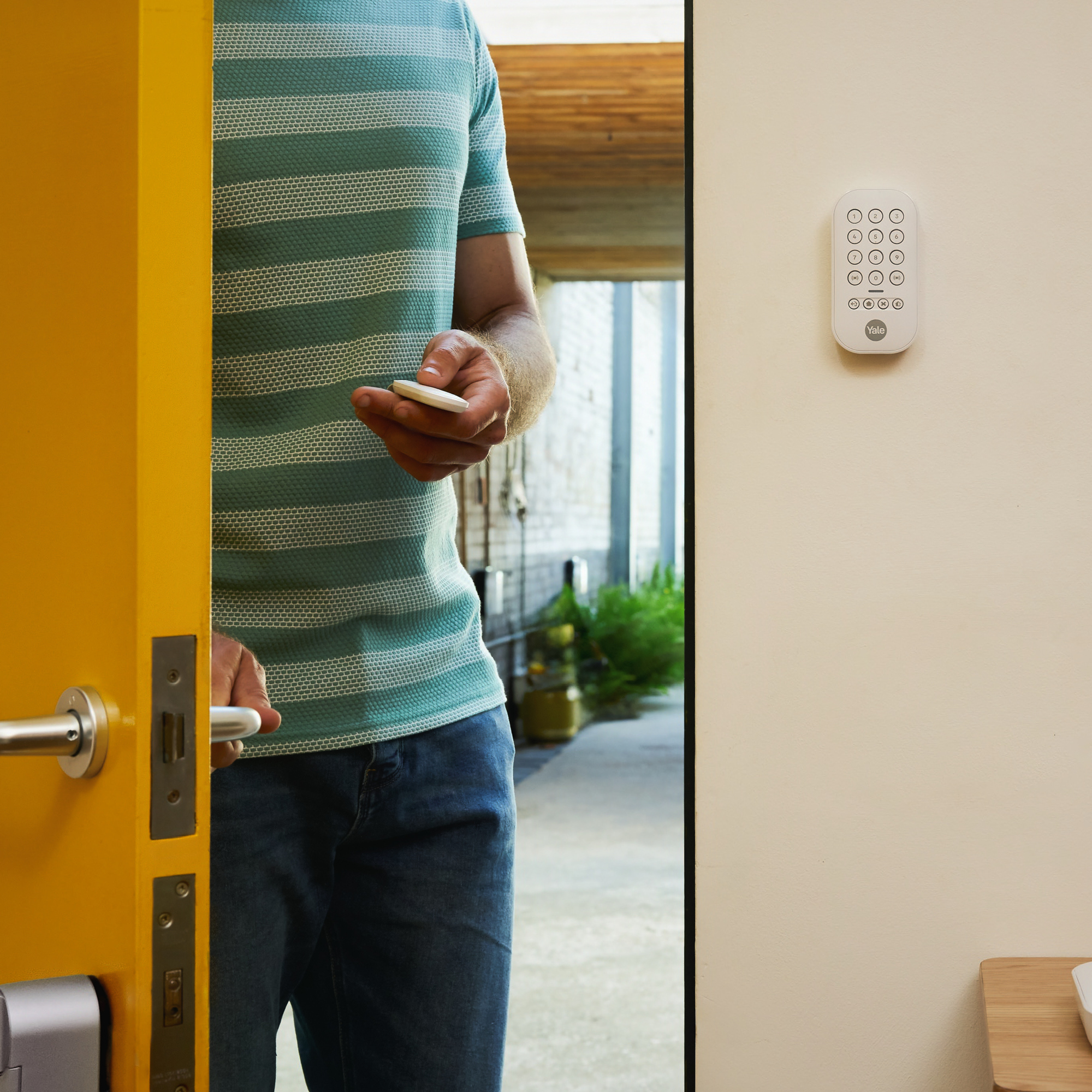 Eine Person hält einen Schlüsselanhänger in der Nähe einer gelben Tür. Ein Sicherheits-Tastaturfeld ist an der Wand montiert. Die Person trägt Jeans und ein gestreiftes Hemd.