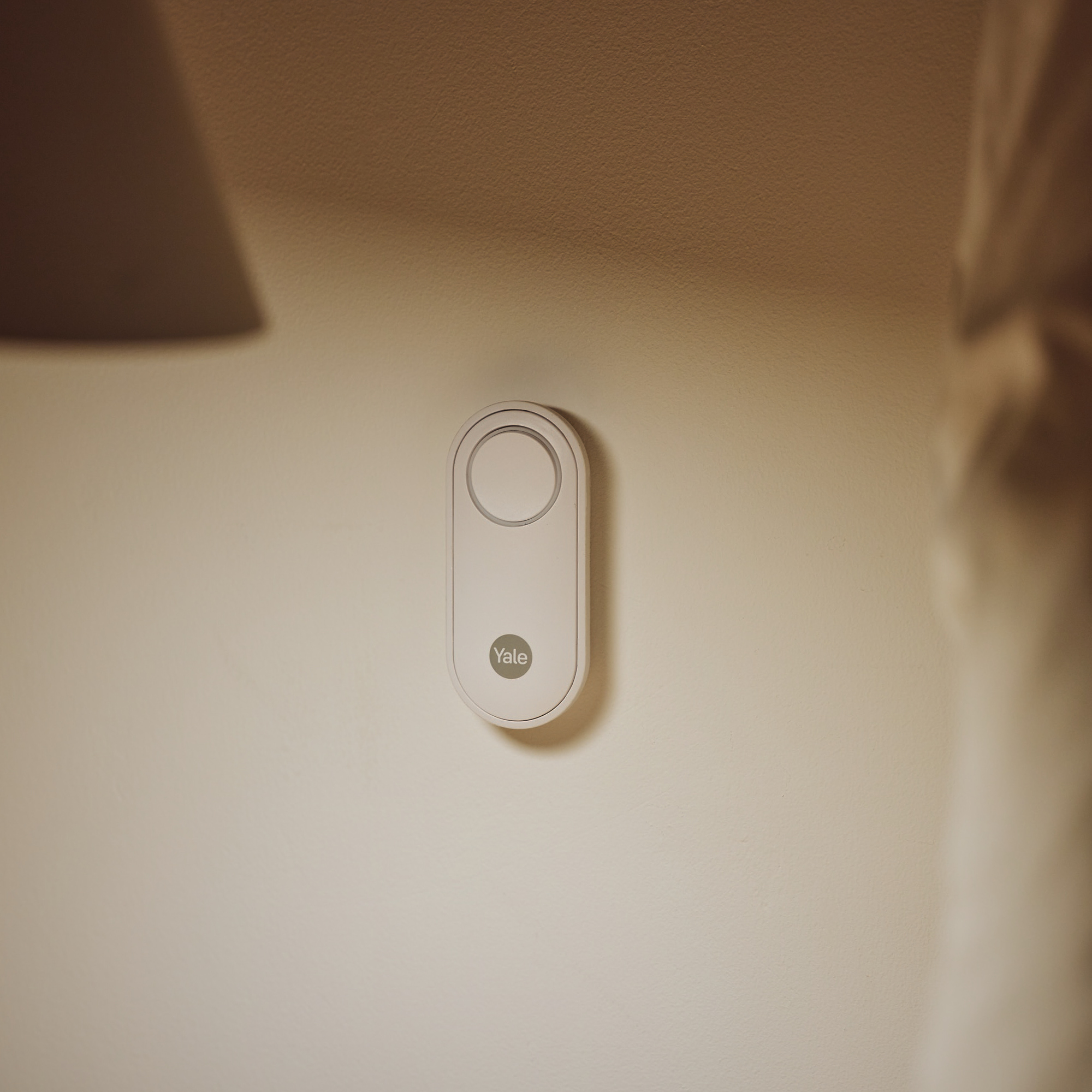 Ein weißer Yale-Türsensor ist an einer hellen Wand montiert. Weiche Schatten werden auf die Wand geworfen.