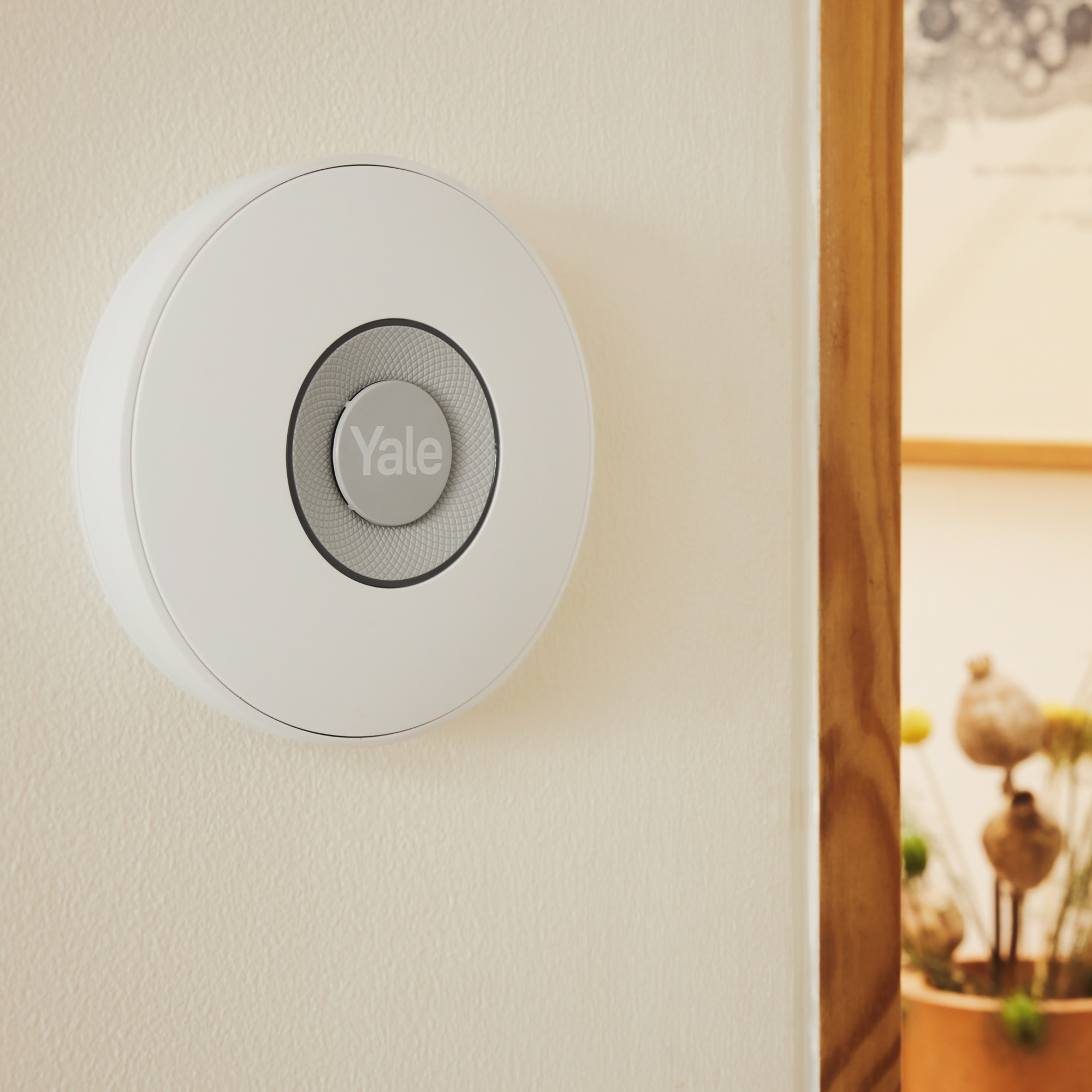 Ein weißes Yale Smart-Home-Gerät ist an einer cremefarbenen Wand montiert. Ein Holzrahmen ist auf der rechten Seite des Bildes sichtbar.