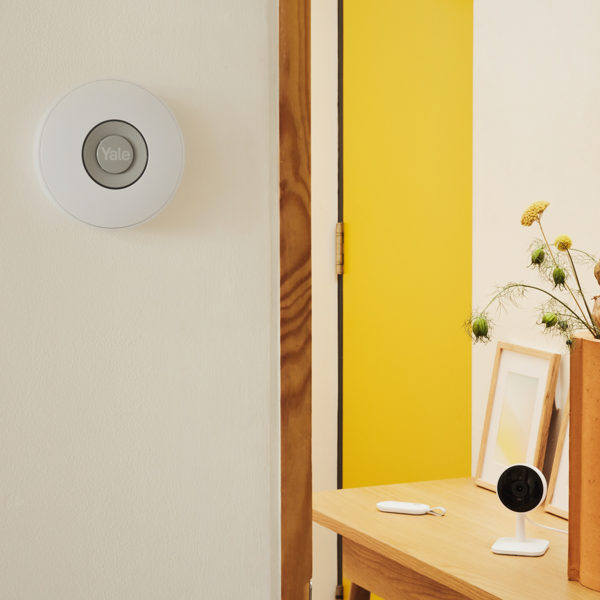 Ein weißes Yale-Sicherheitsgerät an der Wand. Eine gelbe Tür ist geöffnet. Kamera und Deko auf einem Holztisch.