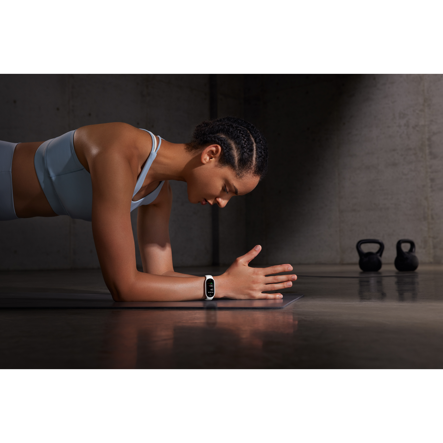 Vrouw in plankpositie met een fitnesstracker. Twee kettlebells op de achtergrond. Draagt een lichtblauwe sportbeha.