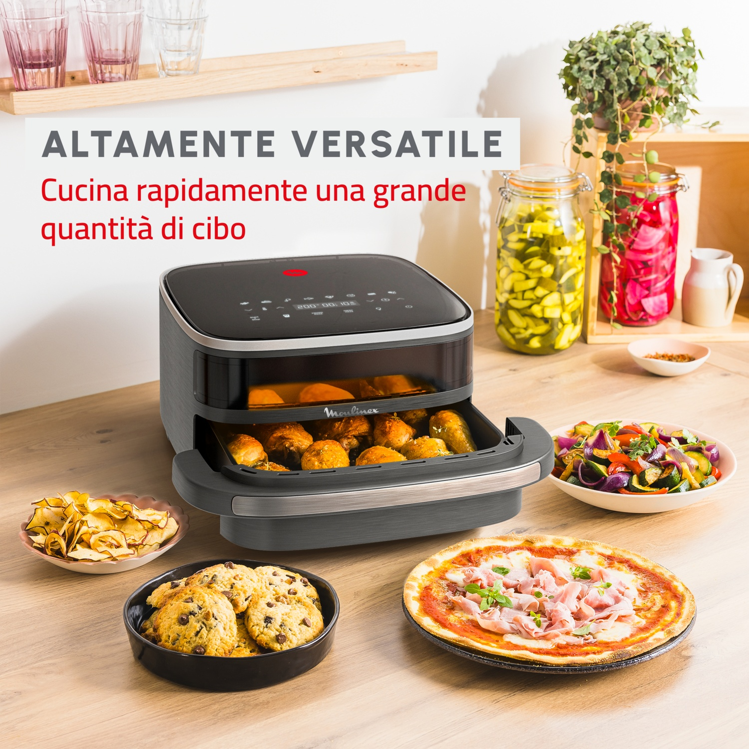Una friggitrice ad aria con cibo, pizza e biscotti su un tavolo di legno. Barattoli di verdure sottaceto sullo sfondo.