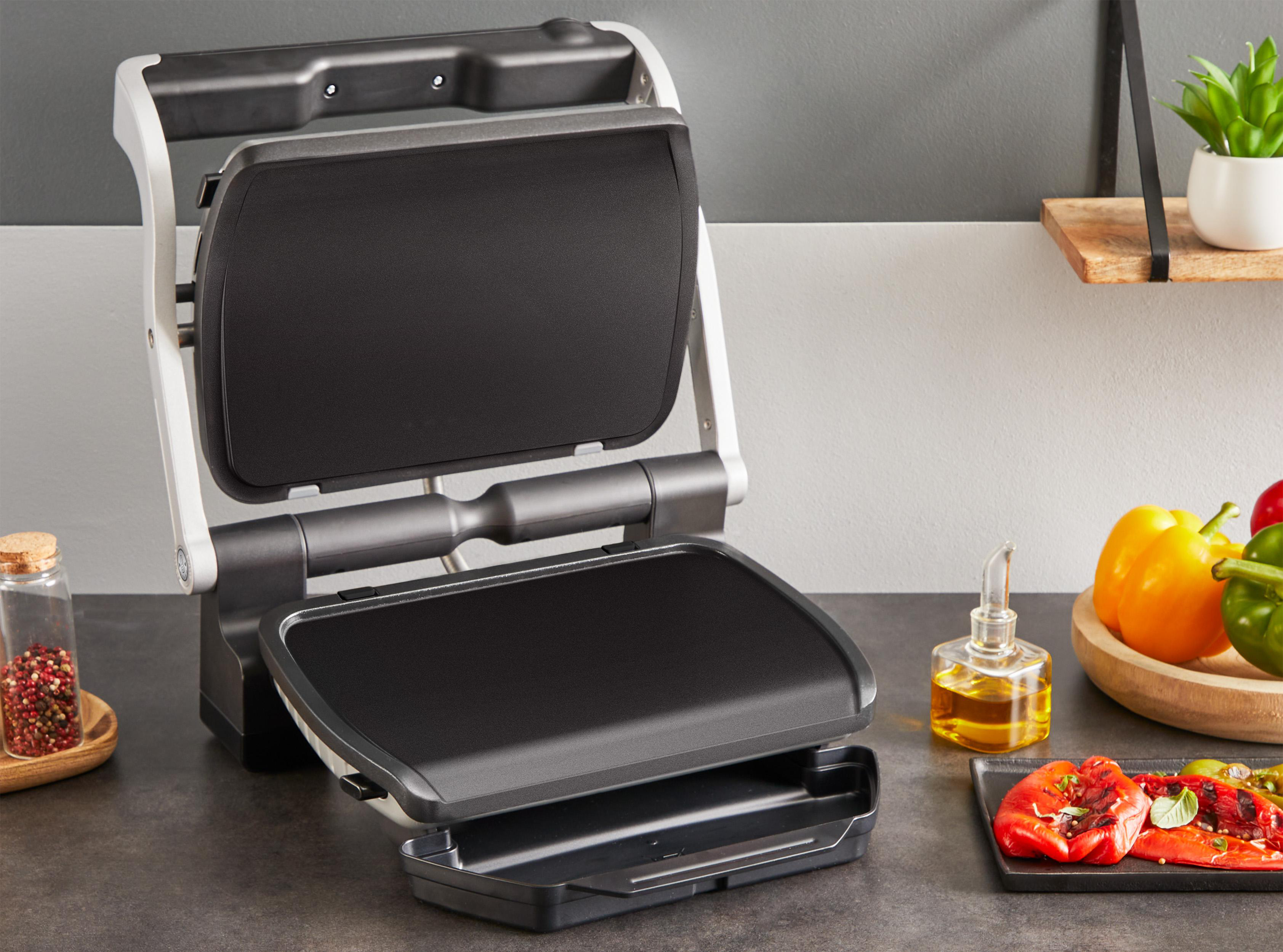 Ein offener Grill mit Gemüse und Öl. Ein Holzregal mit einer Pflanze befindet sich im Hintergrund.