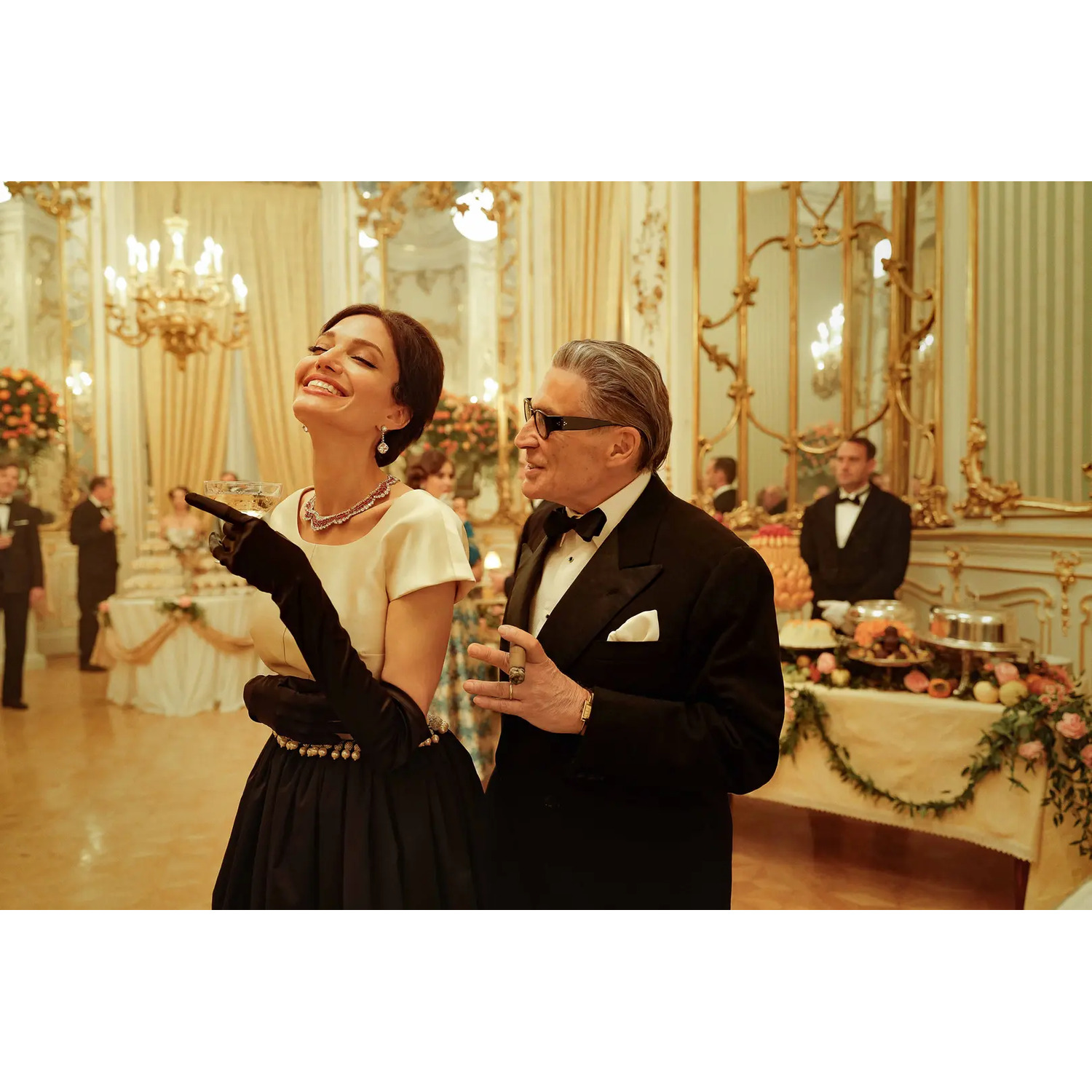 Une femme rit avec un homme en smoking. Ils sont dans une salle de bal avec des lustres et un buffet en arrière-plan.