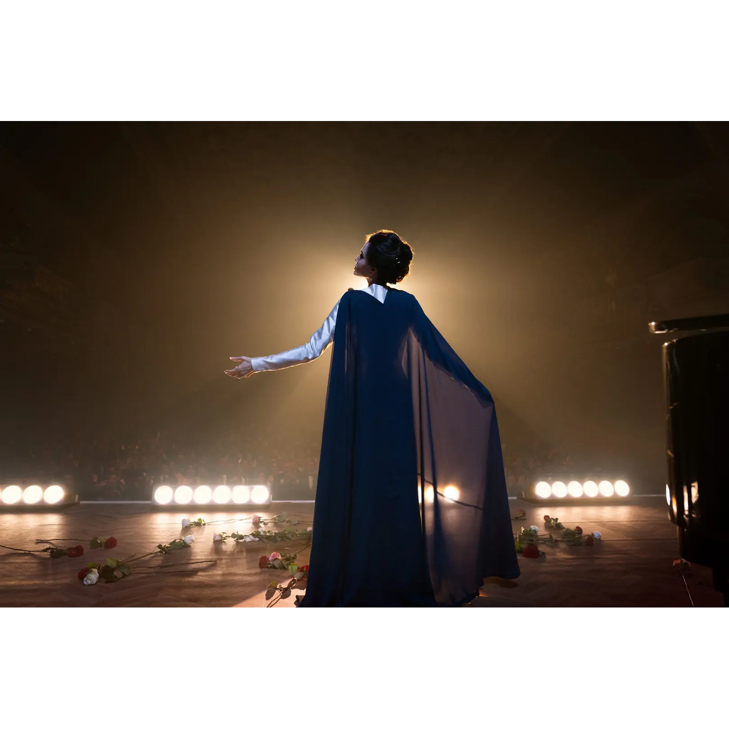Une femme en cape sombre sur scène, bras tendu. Roses éparpillées. Lumières vives brillent de derrière.