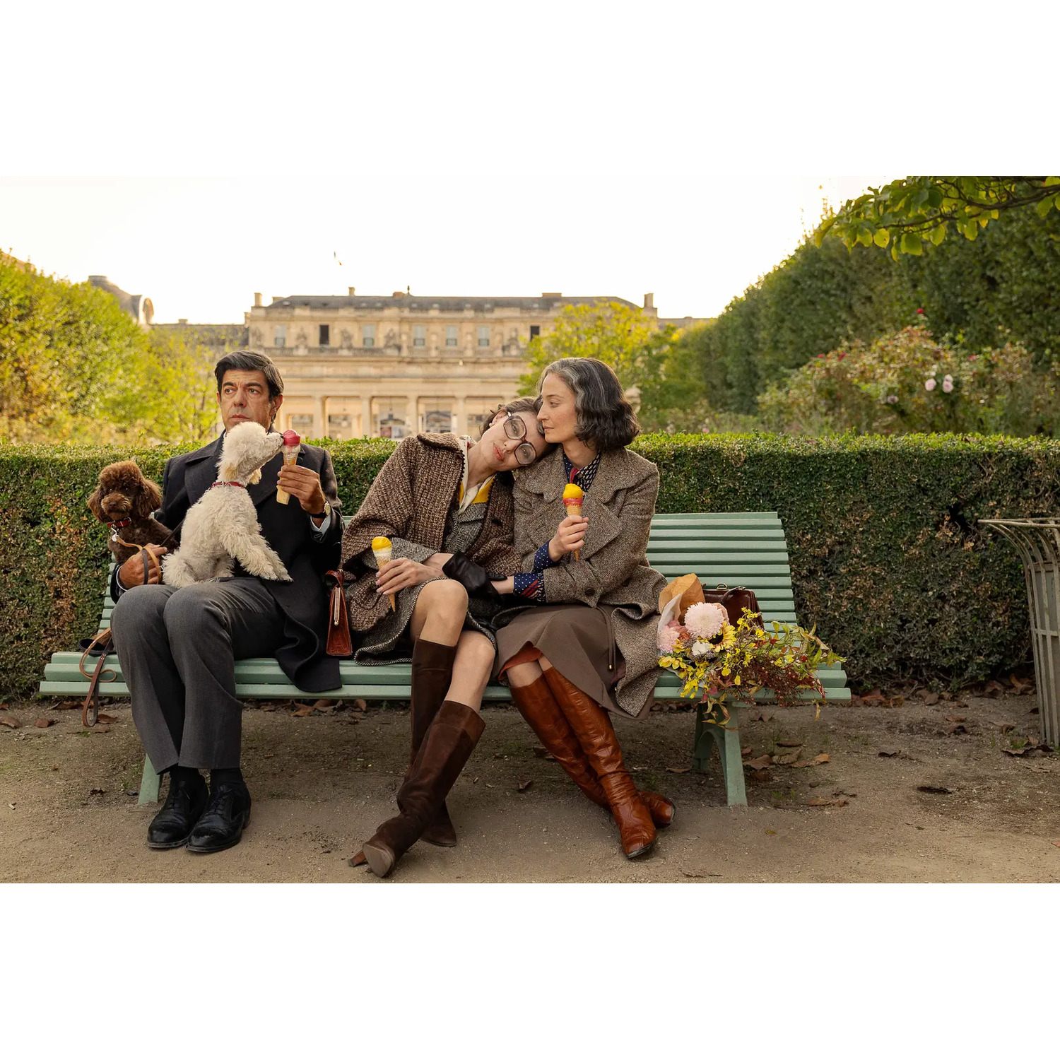 Trois personnes sur un banc dans un parc. Deux chiens et de la glace. Une personne embrasse une autre. Fleurs sur le banc.