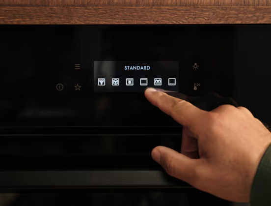 Dłoń dotyka panelu sterowania piekarnika. Ekran wyświetla "STANDARD" i ikony. Piekarnik jest czarny z drewnianym blatem.