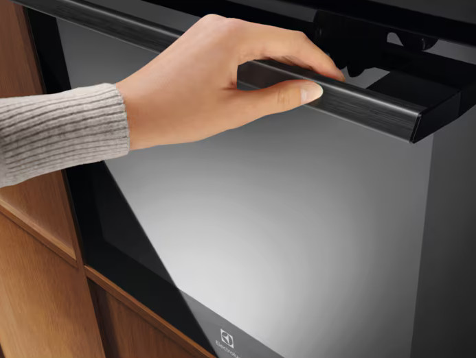 Ręka otwiera czarne drzwi piekarnika. Piekarnik jest czarny, a szafka drewniana. Widoczna jest marka Electrolux.