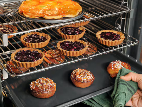 Piekarnik z ciastami i tartami na kratkach. Ręka z zieloną szmatką wyjmuje tacę z cynamonkami.