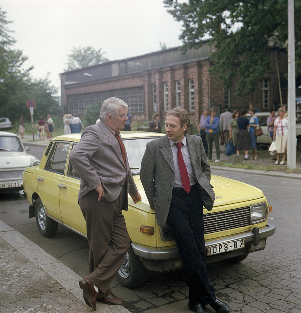 Zwei Männer stehen an einem gelben Auto. Einer lehnt sich an das Auto, der andere steht ihm gegenüber. Im Hintergrund stehen Leute.