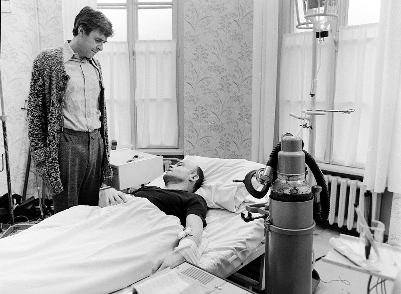Ein Mann in einem Krankenhausbett wird von einem anderen Mann betreut. Medizinische Geräte umgeben das Bett. Das Zimmer hat ein Fenster und Tapeten.