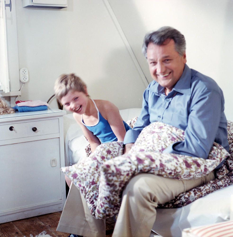 Ein Mann und ein Mädchen lächeln. Der Mann trägt ein blaues Hemd und beige Hose, hält eine geblümte Decke. Das Mädchen trägt ein blaues Oberteil.