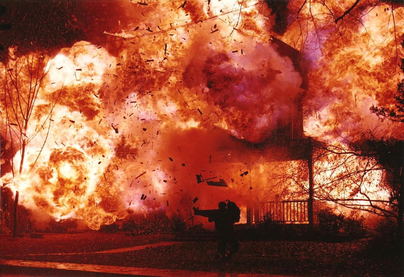 Ein Haus wird von einer Explosion verschlungen. Silhouette einer Person im Vordergrund. Trümmer und Flammen füllen den Himmel.